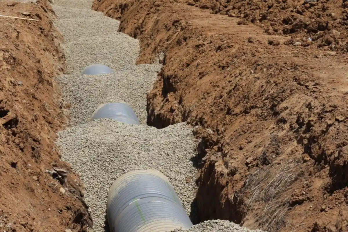 Gray corrugated pipe bedded in gravel at the bottom of a freshly dug dirt trench