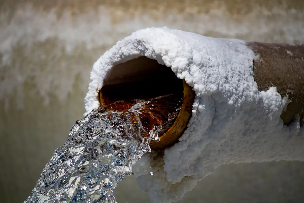 Metal pipe outlet crusted thick with white mineral scale, water pouring out of the rust-stained opening
