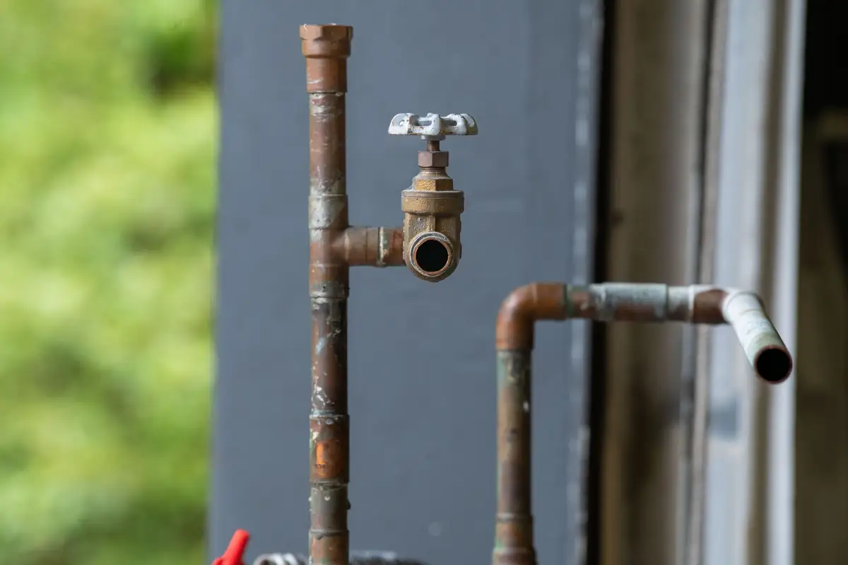 Tarnished, corroded copper pipes with an old brass gate valve outside a gray house wall